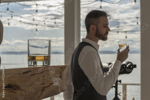 Φωτογραφία Whiskey neat in glass on wood foreground focus with man sipping a glass in backg