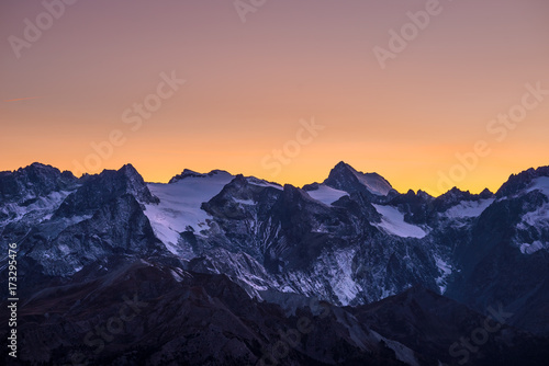 Colorful sky at dusk beyond the glaciers on the majestic peaks of the Massif des Ecrins (4101 m), France. Telephoto view from distant at high altitude. Clear orange sky.