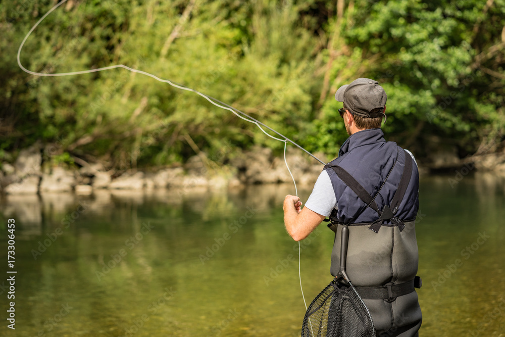 Angler mit Wathose und Fliegenrute im Wasser beim Angeln bei Sonne im klaren Fluss stehend und werfend Angler mit Wathose und Fliegenrute im Wasser beim Angeln bei Sonne im klaren Fluss stehend und werfend
