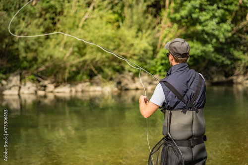 Angler mit Wathose und Fliegenrute im Wasser beim Angeln bei Sonne im klaren Fluss stehend und werfend