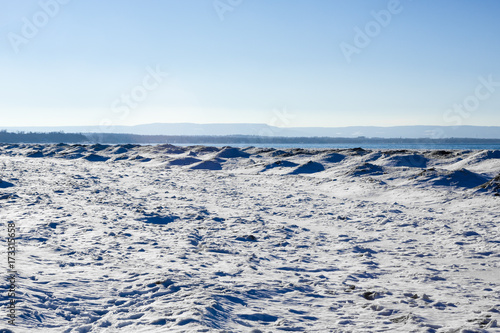 Frozen ice, snow, and sand dunes on beach with hills in distance.