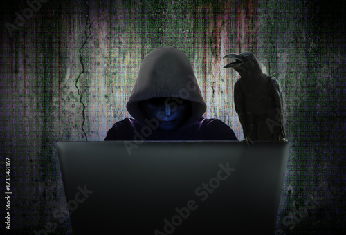 Computer cyber criminals in black mask and hood are hacking data to infiltrate and attack corporate security systems with the black crow is hanging on the computer