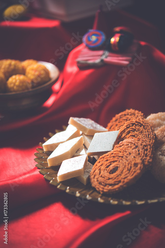 stock photo showing Diwali still life with Diya or oil Lamp, gift boxes, sweets and fire crackers with flowers arranged over red satin cloth with bokeh
 photo