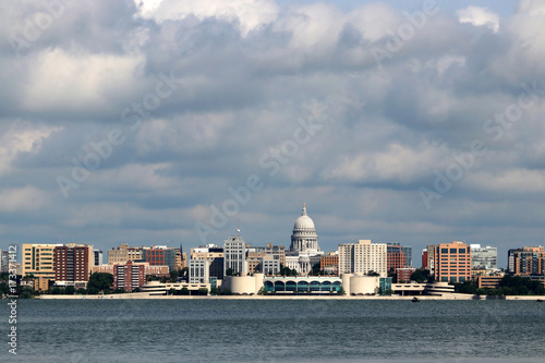 Downtown skyline of Madison, capital city of Wisconsin, USA. Morning view with State Capitol and official buildings in sunlight against beautiful cloudy sky and lake water as seen across lake Monona.