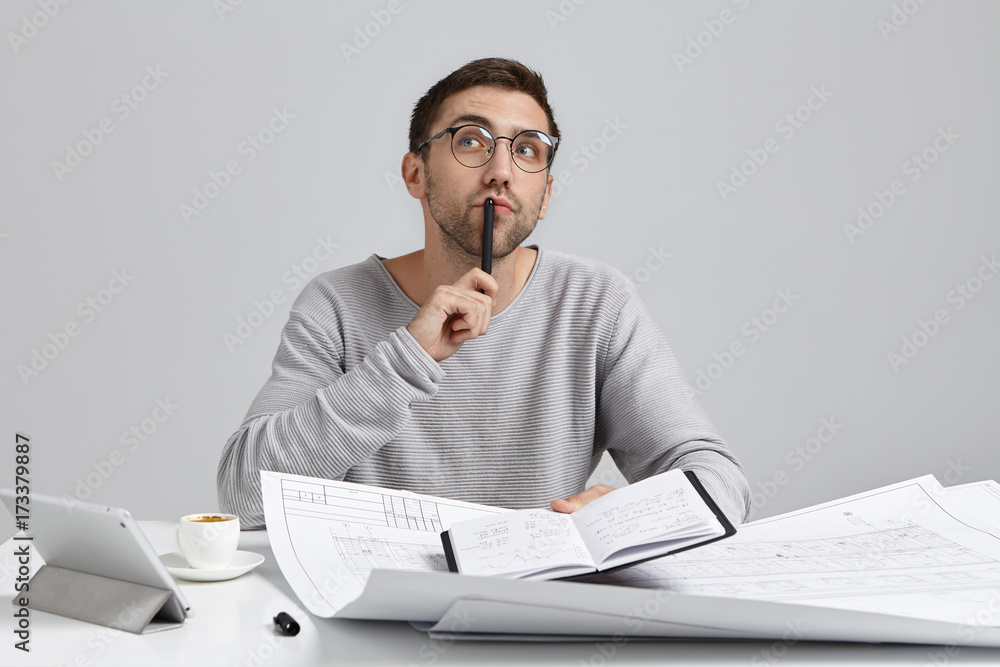 Naklejka premium Waiting for inspiration. Horizontal shot of handsome talented European designer making notes in copybook, looking sideways, holding black pen at his lips, writing down creative ideas and solutions