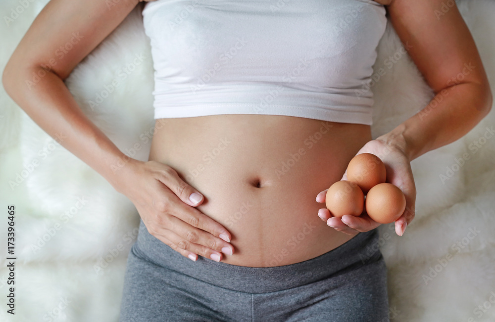 Pregnant woman holding an egg at her belly sitting on the sofa. Pregnant woman holding an egg at her belly sitting on the sofa.