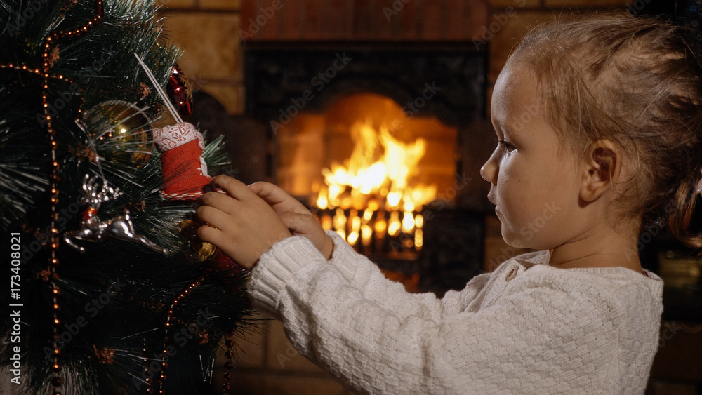 Cute little girl decorating Christmas tree in dark room with fireplace