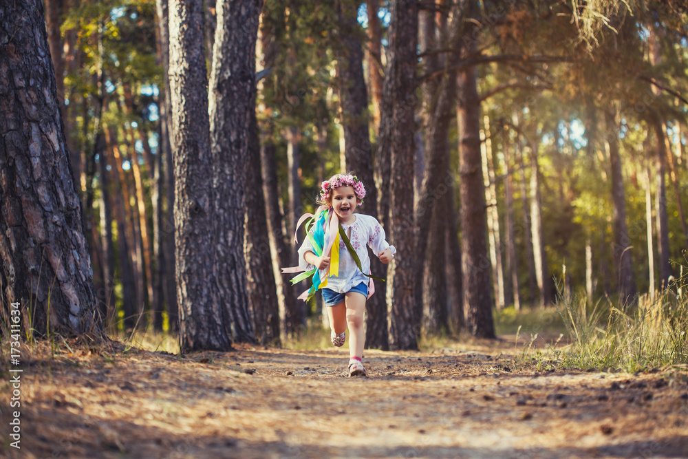 A little girl in a wreath with ribbons is running happily through the summer forest. A little girl in a wreath with ribbons is running happily through the summer forest.