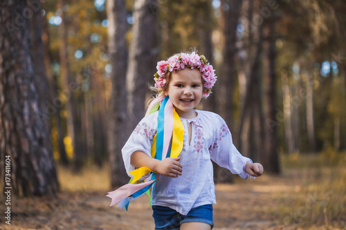 A little girl in a wreath with ribbons is running happily through the summer forest. © Dmytro Titov