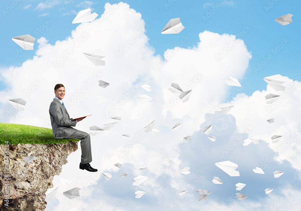 Man on edge reading book and paper planes flying in air Man on edge reading book and paper planes flying in air