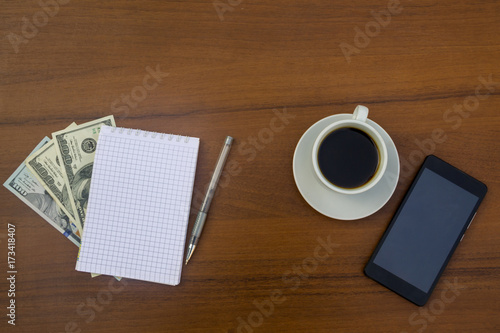 Cup of coffee, smartphone, dollars, notepad and pen on wooden desk