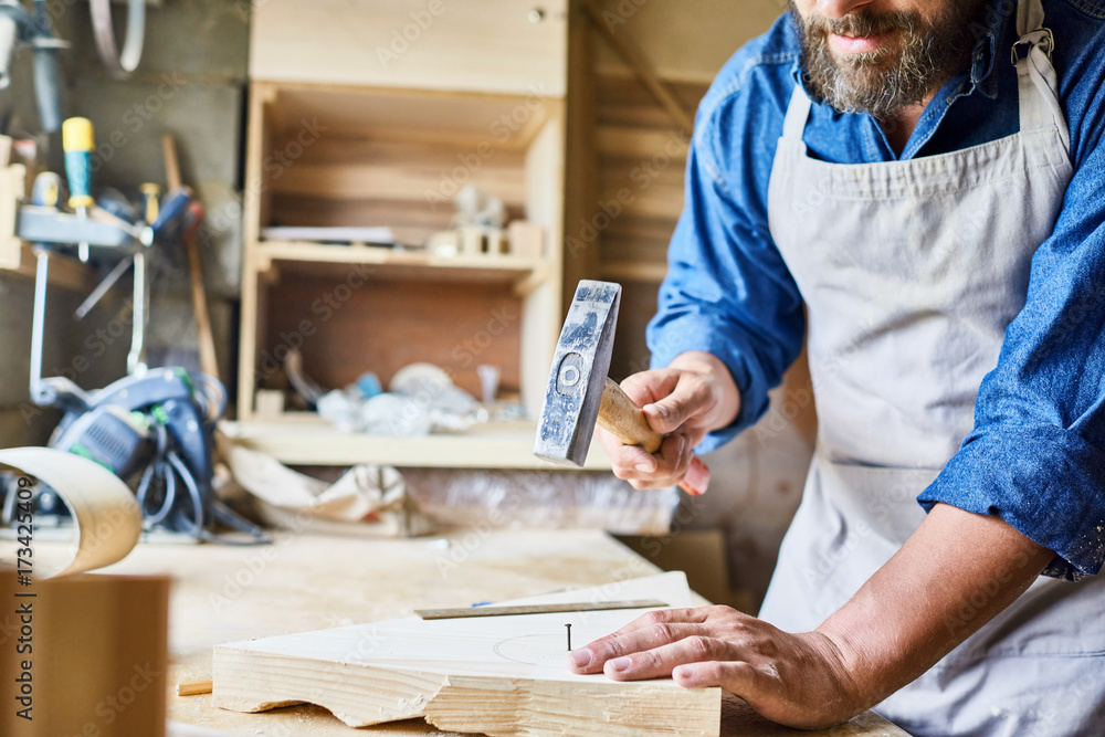 Closeup portrait of skilled carpenter making wooden furniture in workshop hammering nail in piece of wood, copy space Closeup portrait of skilled carpenter making wooden furniture in workshop hammering nail in piece of wood, copy space