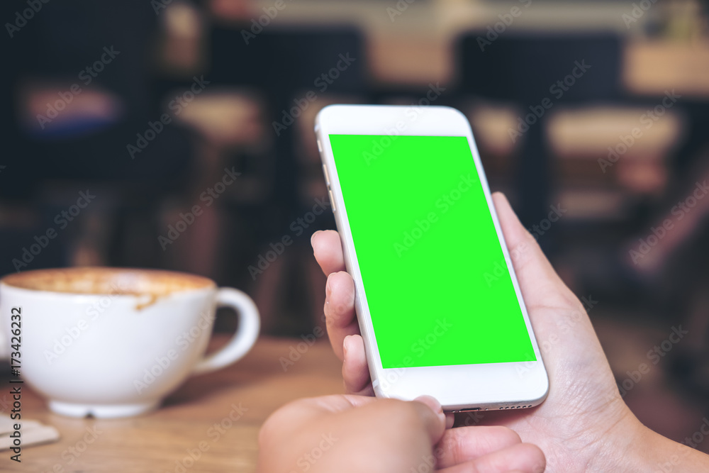 Mockup image of hands holding white mobile phone with blank green screen with coffee cups on wooden table in restaurant