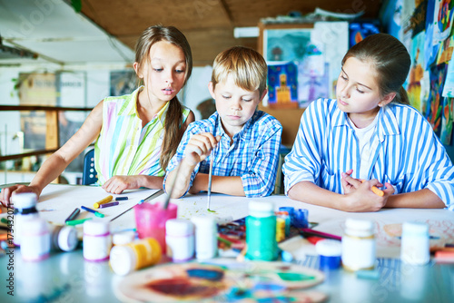 Group of three children  two girls and one boy   painting together in art class sitting at big table with art supplies  pencils and paints