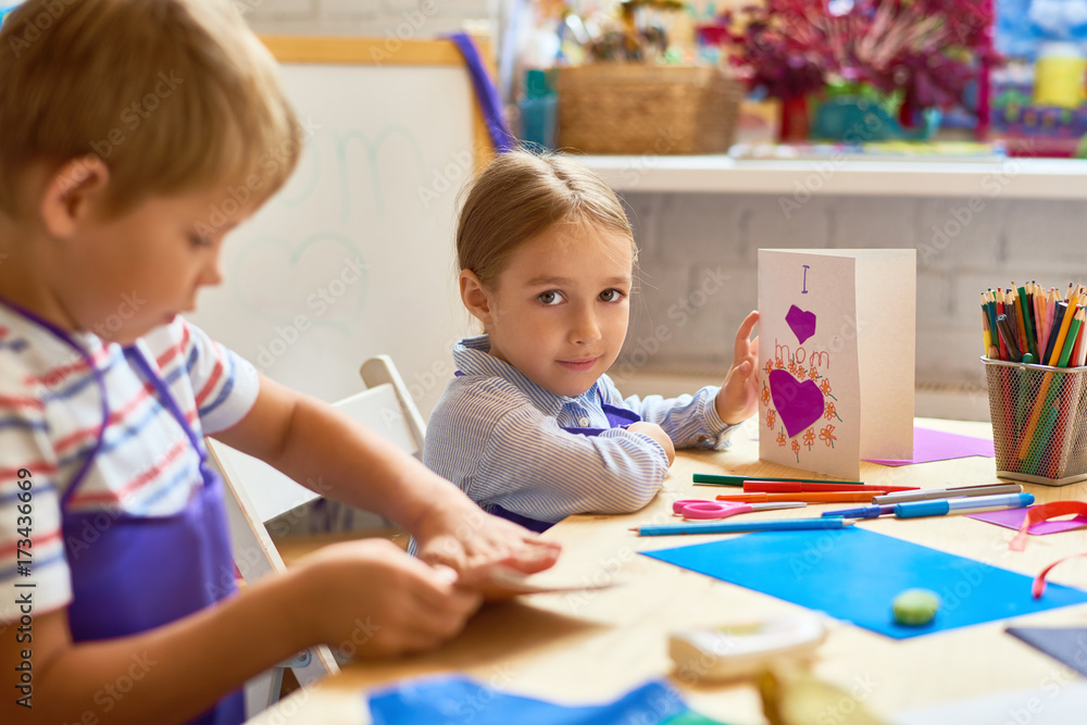 Portrait of cute little girl holding handmade gift card for mom and looking at camera while sitting at desk during art class in pre-school Portrait of cute little girl holding handmade gift card for mom and looking at camera while sitting at desk during art class in pre-school