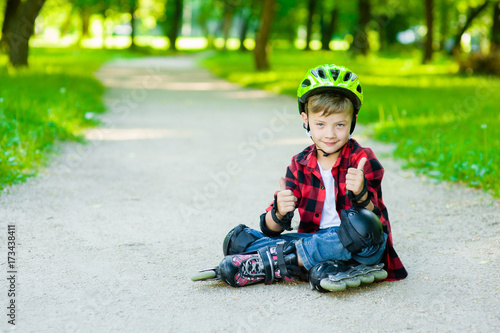 Boy in a protective helmet and protective pads for roller skating sitting on the road and showing thumbs up © Ermolaev Alexandr