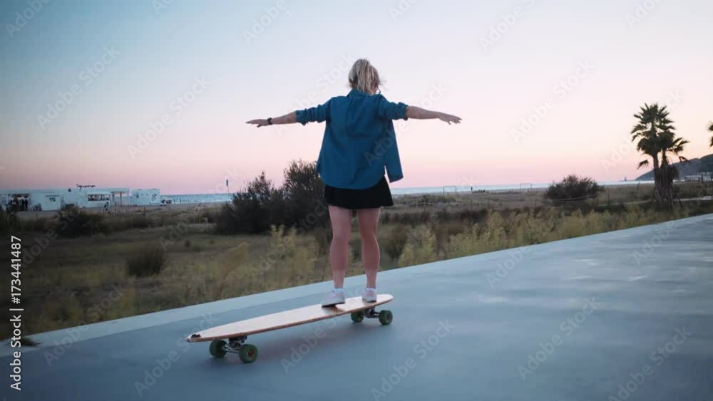 Beautiful soft pink colors of pastel sunset in last days of summer. young hipster woman or teenager rides skateboard or longboard next to emppty beach promenade