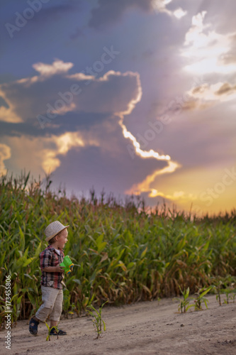 A boy in a hat is walking at sunset. A small child in the background of a cornfield.
