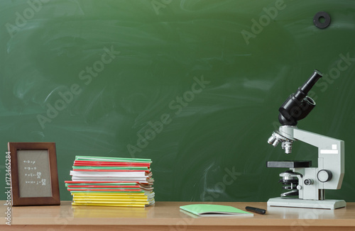 Teacher or student desk table. Education background. Education concept. Copybook, books, pen, microscope and photo frame on the table on blackboard (chalkboard). Chemistry or biology lesson.