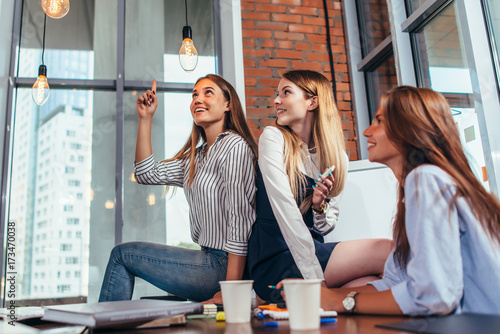 Fotografi Happy female teenagers sitting on table looking and pointing at stylish light bu