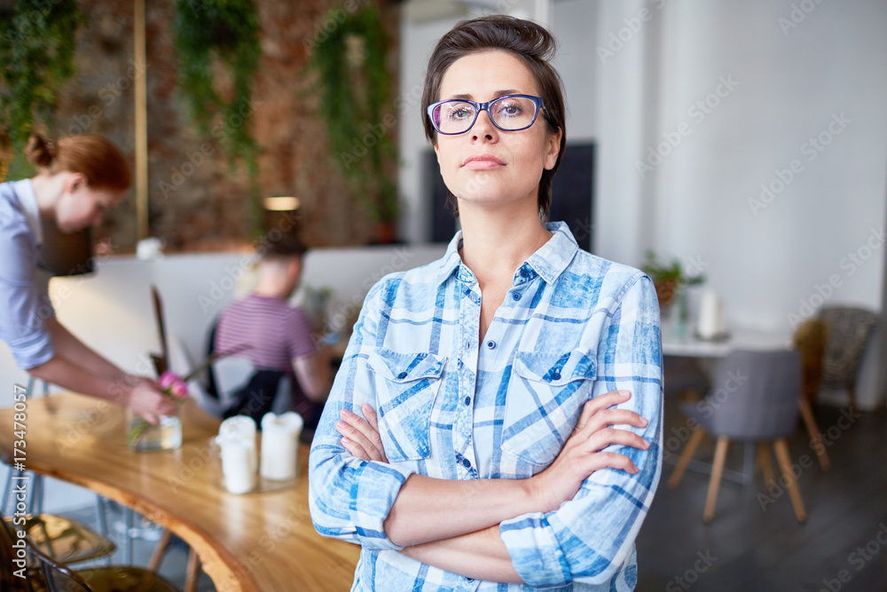 Confident owner of cafe crossing her arms on chest with waitress working on background