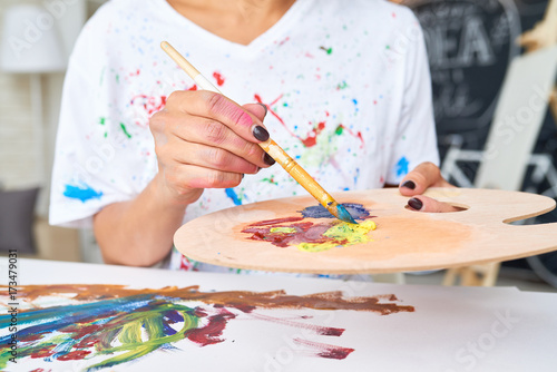 Closeup of young woman mixing oil colors on palette with big paintbrush while enjoying hobby in modern apartment