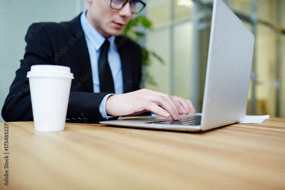 Close-up shot of confident young manager wearing suit sitting in front of laptop and writing response email to client, interior of spacious open plan office on background Close-up shot of confident young manager wearing suit sitting in front of laptop and writing response email to client, interior of spacious open plan office on background