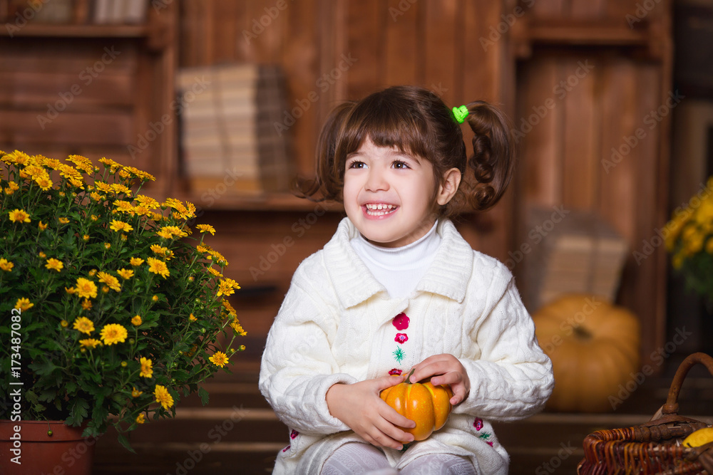 Fototapeta premium Portrait of adorable smiling girl posing with orange pumpkin in fall wooden interior.