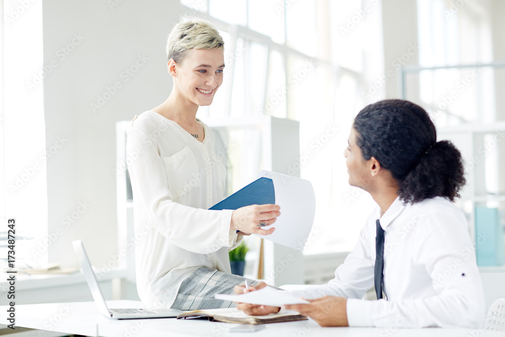 Pretty young financial manager with charming smile preparing annual accounts with handsome mixed race colleague while gathered together at modern open plan office Pretty young financial manager with charming smile preparing annual accounts with handsome mixed race colleague while gathered together at modern open plan office