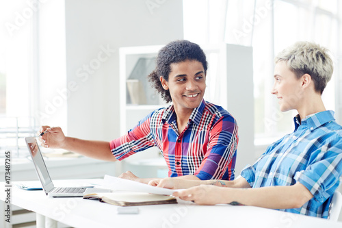 Smiling young interior designers wearing checked shirts working together on joint project while sitting at desk in spacious open plan office, handsome mixed race man pointing at laptop screen