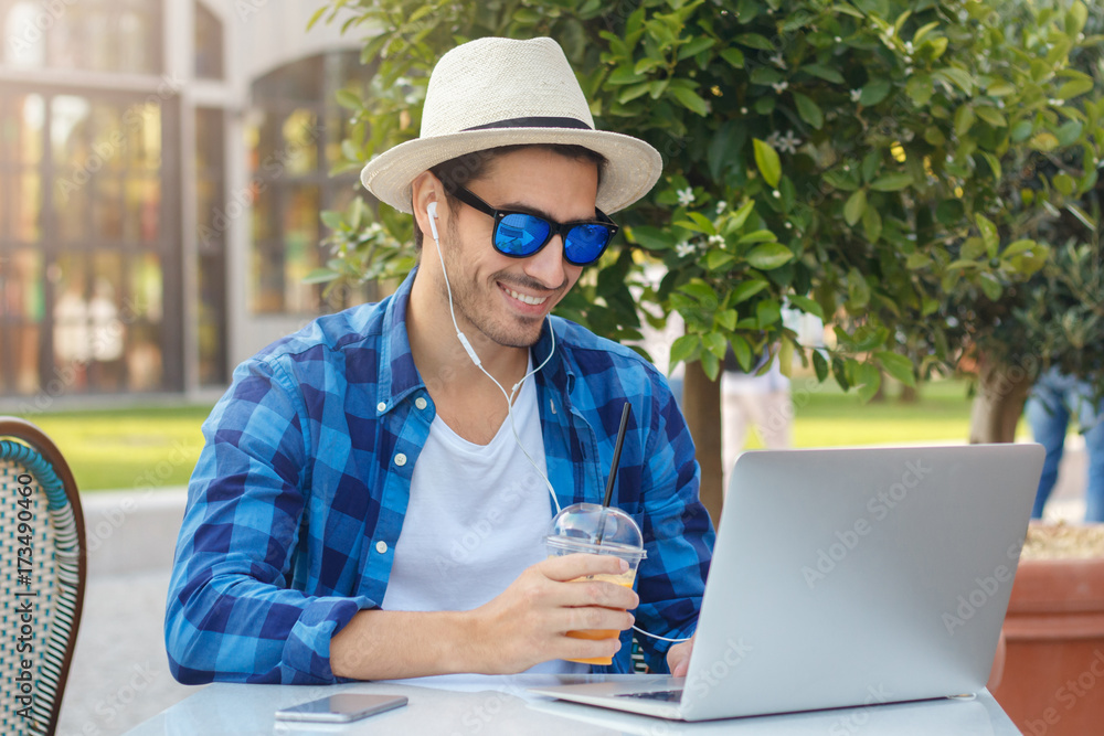 Outdoor photo of positive European guy enjoying free time at cafe table while checking social networks via laptop, browsing and listening to music or answering video call with happy friendly smile