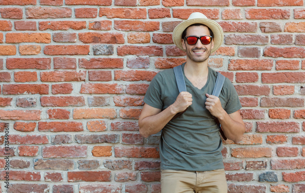 Outdoor portrait of young Caucasian man pictured with red brick wall in background looking through sunglasses with joyful smile, pulling straps of backpack forward as if ready to continue journey Outdoor portrait of young Caucasian man pictured with red brick wall in background looking through sunglasses with joyful smile, pulling straps of backpack forward as if ready to continue journey