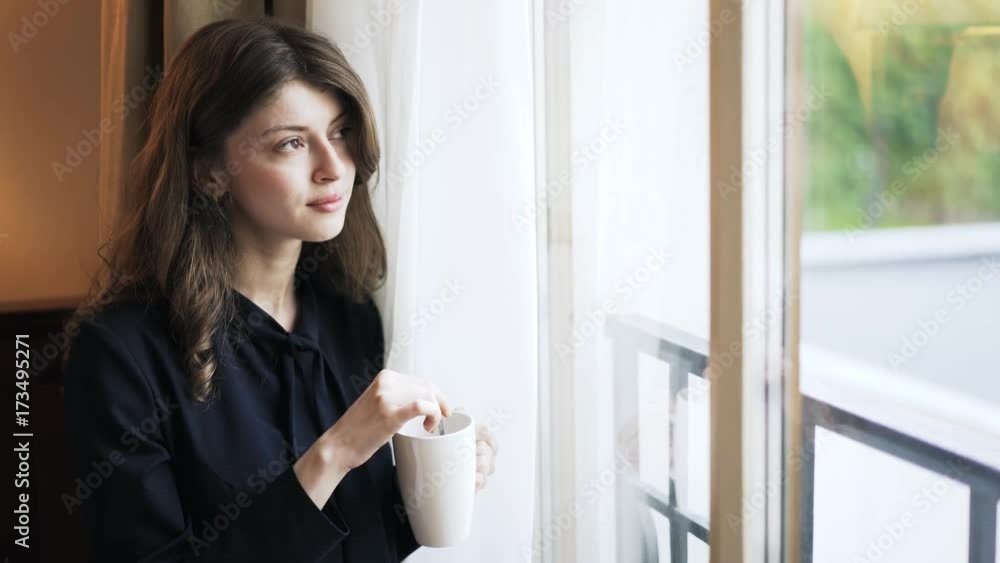 Pensive young woman wearing black clothes is holding a white mug and looking in a window. Handheld real time close up shot Pensive young woman wearing black clothes is holding a white mug and looking in a window. Handheld real time close up shot