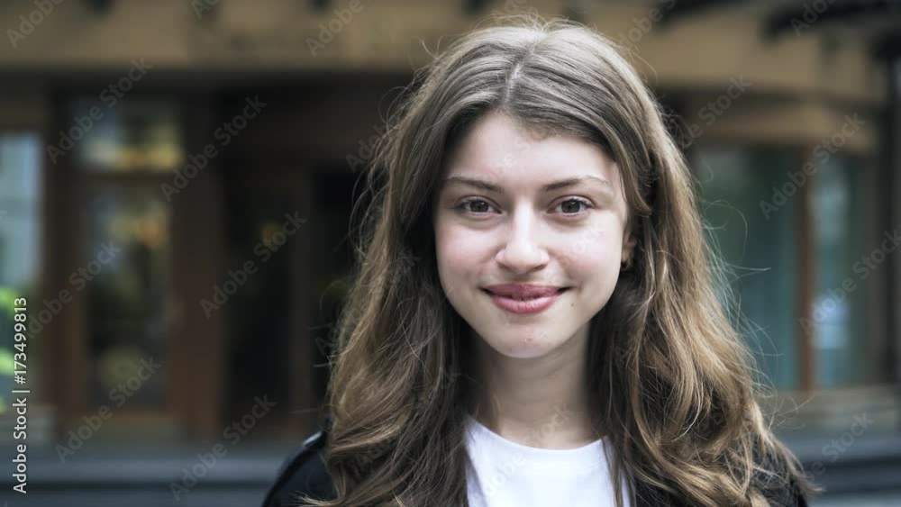 Close up of a beautiful young woman with long brown hair looking at the viewer and smiling while standing in the street on a summer day. Handheld real time close up shot Close up of a beautiful young woman with long brown hair looking at the viewer and smiling while standing in the street on a summer day. Handheld real time close up shot