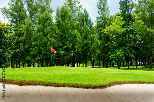 beautiful idyllic view of green with sand trap and red flag and view of forest, blue sky with clouds