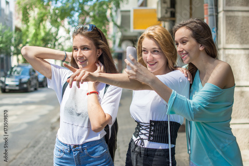 Three happy teenage girls looking something in front of them and taking a photograph on mobile phone, outdoors.