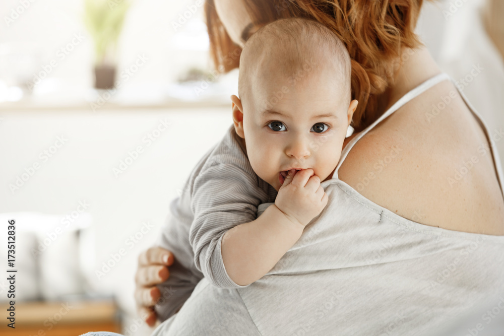 Portrait of sweet little child looking at camera with big grey eyes and putting hand in mouth on mother shoulder. Mom hug her child with love. Family concept. Portrait of sweet little child looking at camera with big grey eyes and putting hand in mouth on mother shoulder. Mom hug her child with love. Family concept.