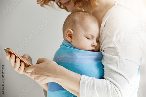 Bild auf Leinwand Beautiful young mother reads articles about maternity life on phone while little lovely son sleeping on her chest in blue baby sling