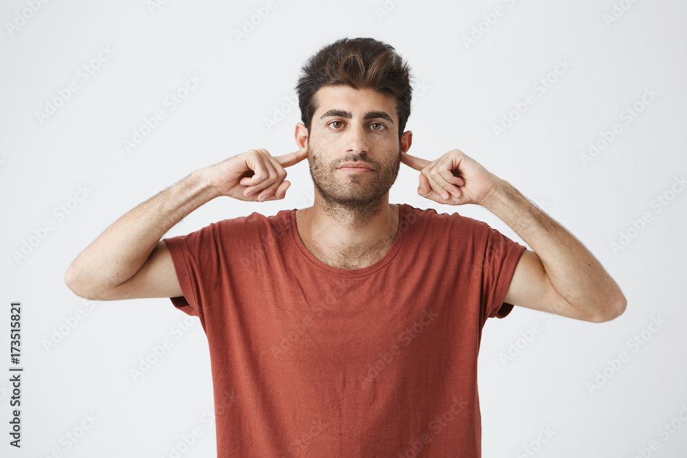 Headshot of annoyed stylish italian guy in red t shirt looking grumpy, closing ears with fingers showing disrespect on his friend words. Headshot of annoyed stylish italian guy in red t shirt looking grumpy, closing ears with fingers showing disrespect on his friend words.