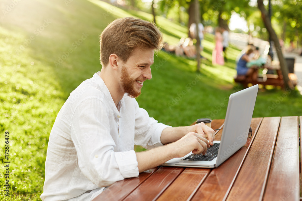 Portrait of attractive red-head man with beard in white shirt, sitting in the park, laughing and doing his job on laptop. Freelance concept. Portrait of attractive red-head man with beard in white shirt, sitting in the park, laughing and doing his job on laptop. Freelance concept.