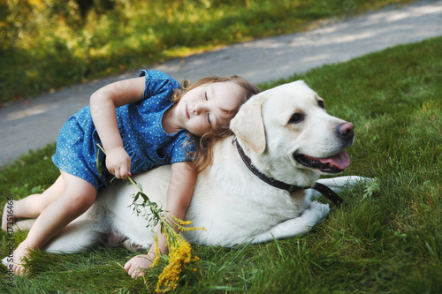Little girl sitting on the grass with labrador retriever in the summer park