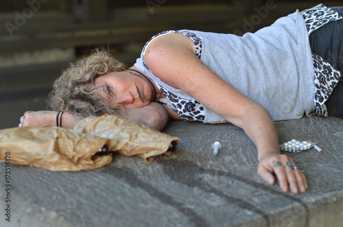 a drunk, homeless woman sleeping under a bridge with bottles of alcohol and other drugs around her