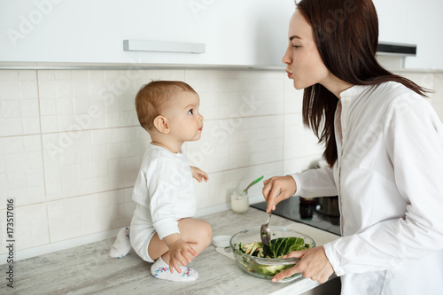 Obraz na plátně Young beautiful mother cooking salad for breakfast and trying to kiss her little son while he looking at mom with interested expression