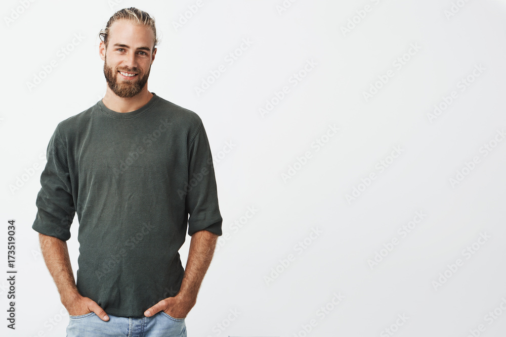 Handsome nordic man with beard and stylish hairstyle in grey shirt and jeans smiling, looking in camera, keeps hands in pockets. Handsome nordic man with beard and stylish hairstyle in grey shirt and jeans smiling, looking in camera, keeps hands in pockets.