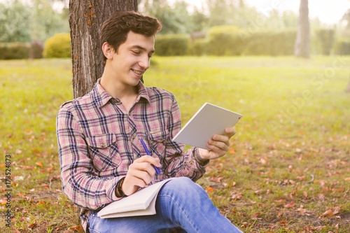 young men student sitting in the park next to the tree with the tablet in his hands