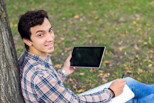 young men student sitting  in the park next to the tree with the tablet in his hands