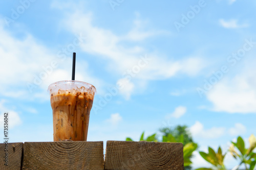 close up of iced latte coffee in transparent plastic glass and straw with on the wooden table with blue sky and cloud. coffee and cafe concept.