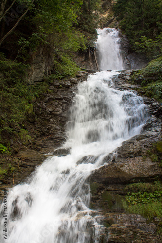 Mountain waterfall in a forest in Valle d'Aosta, flows between trees and rocks.