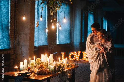beautiful couple embracing, standing near a decorated wooden table for dinner for two, with candles, plates, flowers, light bulbs in a dark loft style room.