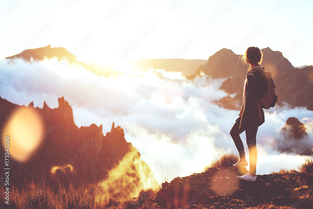 Young energetic girl with  backpack standing at edge of cliff in mountains and looking at sunset with a clouds. - obrazy, fototapety, plakaty 